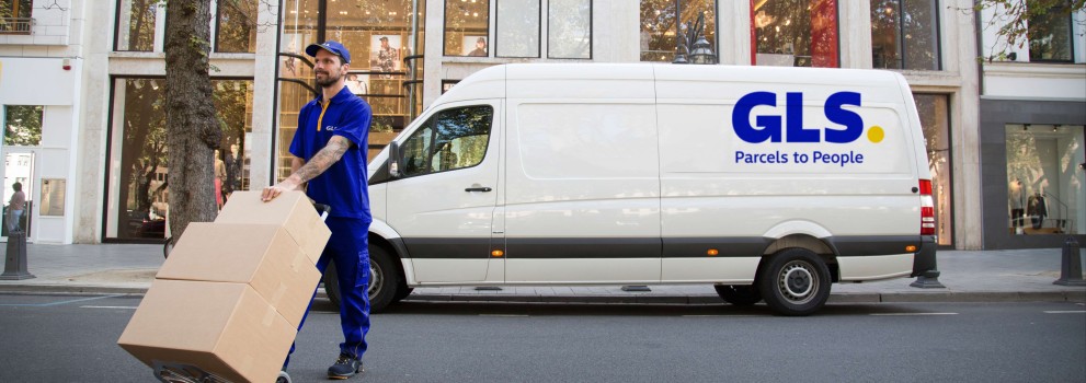 A GLS driver crosses the street, moving packages on a dolly in front of a GLS van.