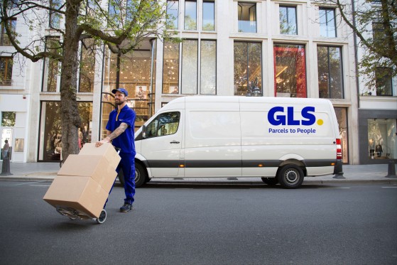 A delivery driver crossing the street, moving packages on a dolly in front of a GLS van.